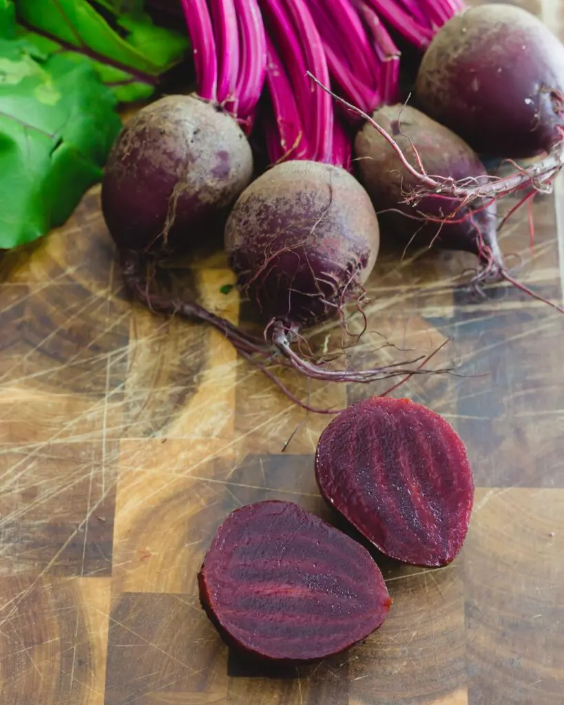 Cooked red beet cut in half on a cutting board with raw beets and beet greens in the background.