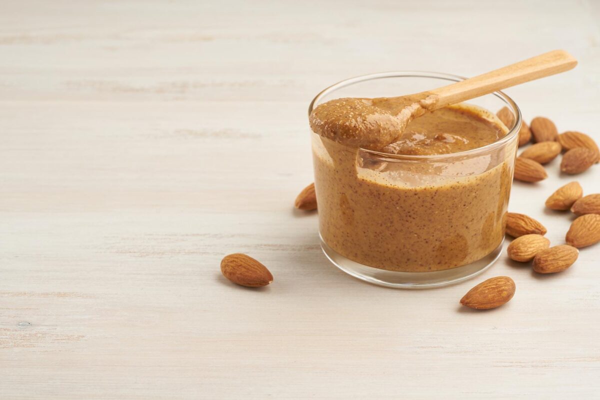 Drippy almond butter in a small glass with wooden spoon and almonds surrounding it.