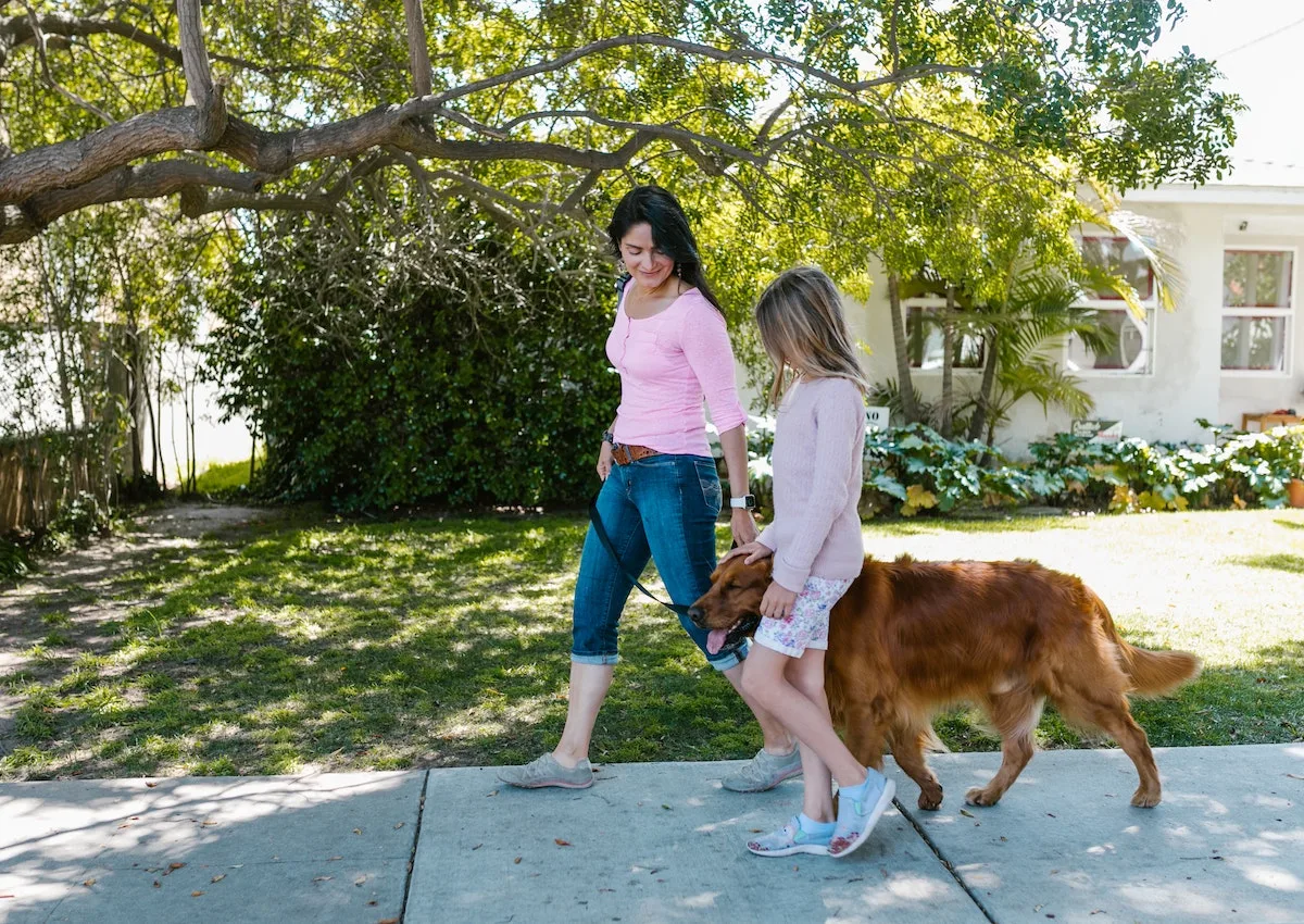 A woman and her daughter walking a dog on a sidewalk.