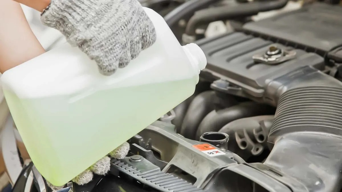 A person wearing gloves is pouring liquid from a white jug into a car engine.