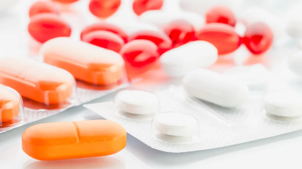 Various prescription pills and capsules in red, white, and orange, arranged on a reflective white surface.