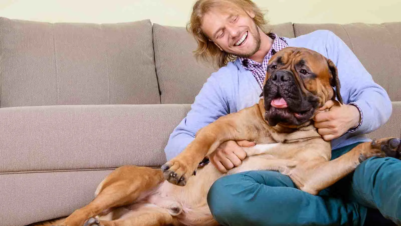A man with long hair sits on the floor against a sofa, smiling and holding a large dog that appears relaxed and happy.