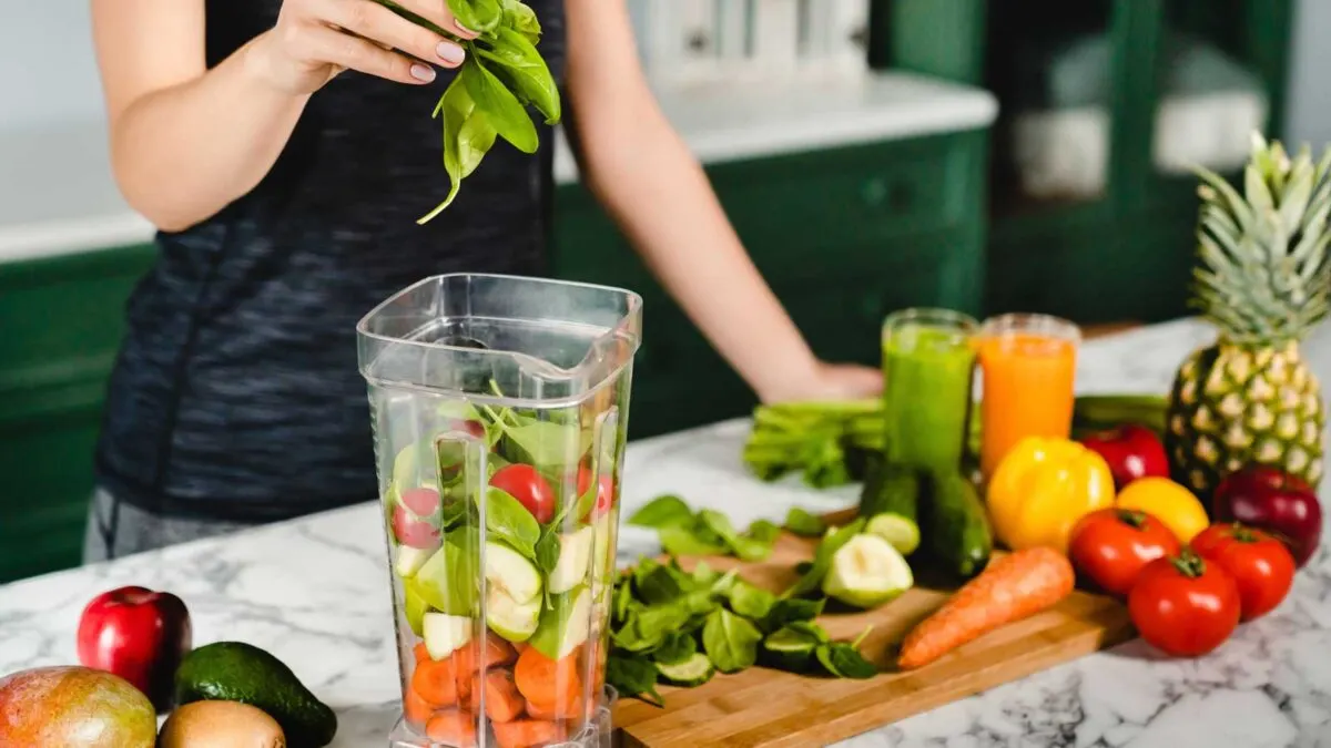 A person adding fresh spinach to a blender filled with various fruits and vegetables on a kitchen counter.