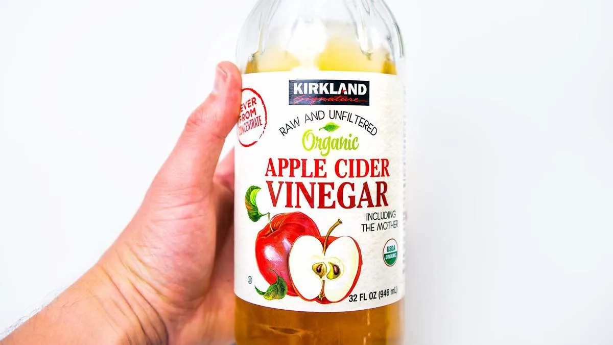 A hand holding a 32-ounce bottle of Kirkland Signature organic raw and unfiltered apple cider vinegar. The label features images of red and sliced apples and notes it includes "the mother.