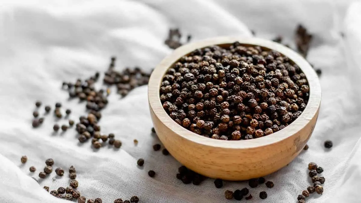 A wooden bowl filled with whole black peppercorns sits on a white cloth surface. Some peppercorns are scattered around the bowl.