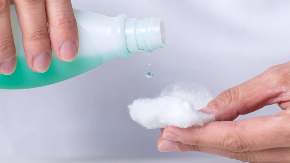 A hand is pouring liquid from a green bottle onto a piece of white cotton held by another hand. The background is softly blurred, emphasizing the focus on the hands and bottle.