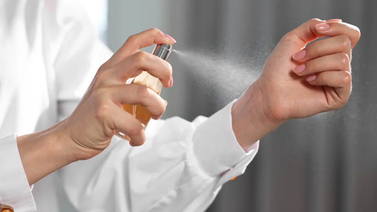 A person sprays perfume on their wrist. They are wearing a white long-sleeved shirt. The background is softly blurred.