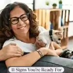 A smiling older woman with glasses sits on a couch holding a small dog—possibly one of the beloved Italian dog breeds—with the text “11 Signs You’re Ready for a Furry Sidekick in Retirement” displayed at the bottom.