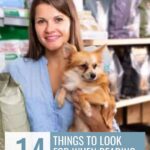 Woman holding a small dog in a pet store, smiling, with shelves of pet products behind her. Text reads &ldquo;14 things to look for when reading dog food labels.&rdquo; Learn how to read dog food labels effectively to make the best choice for your furry friend.